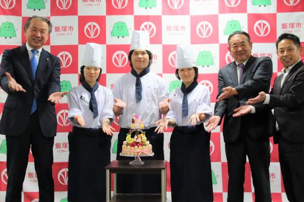 決戦で作成するケーキを前にポージングする武井市長、飯塚高校製菓部3人、学校関係者