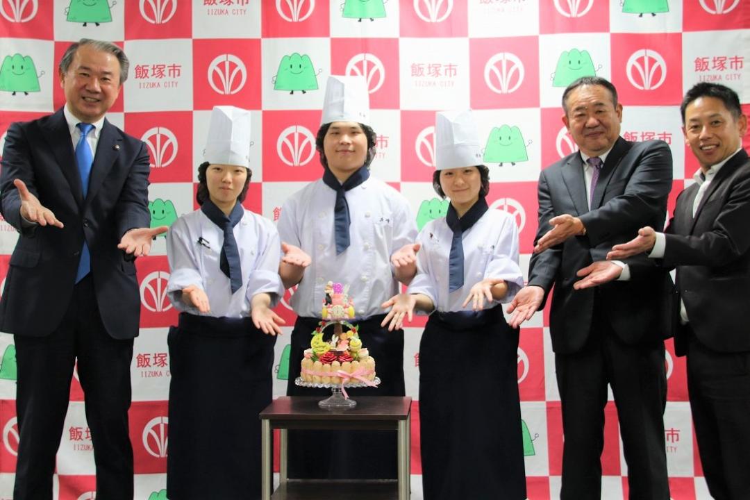 決戦で作成するケーキを前にポージングする武井市長、飯塚高校製菓部3人、学校関係者